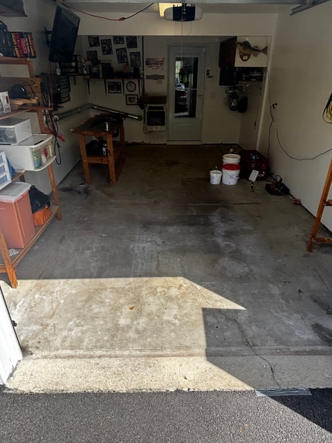 Interior of a garage workshop with concrete floor, workbench, tools on walls, storage cabinets, and open garage door showing ramp